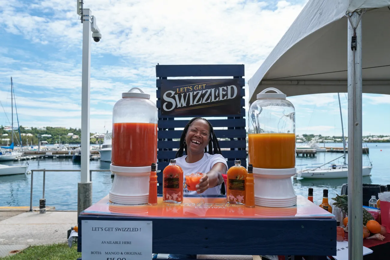 A woman is standing at a bar handing to the camera a rum swizzle. 