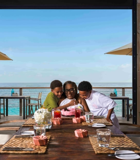 A woman and two children are celebrating a birthday a table with an ocean view in the background.