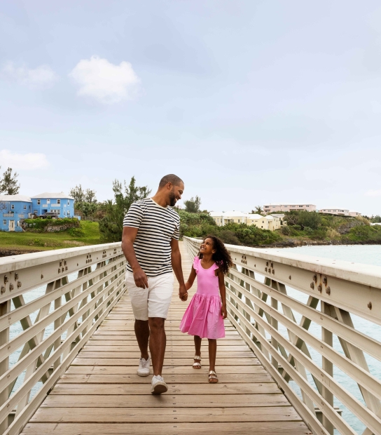 A father and daughter are walking along a bridge smiling.
