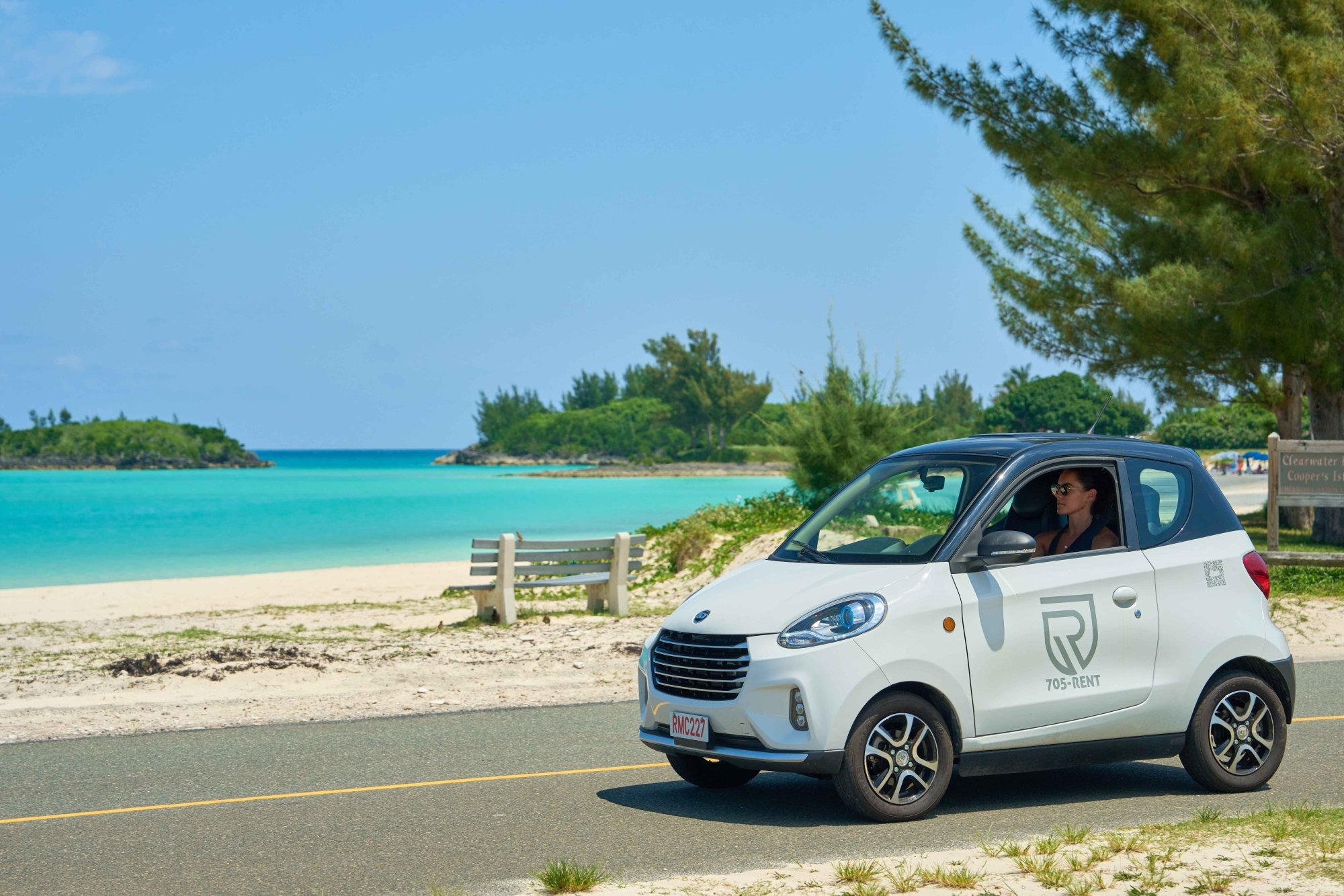 A woman is driving a micro car with a beach in the background.